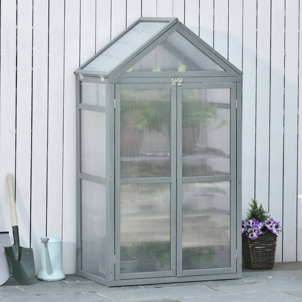 Serre grise fermée et garnie de plantes devant une palissade blanche